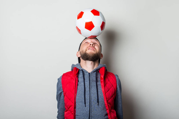 Young man in a cap, hoodie and vest holds a soccer ball on his head on a light background. - Foto, Imagen