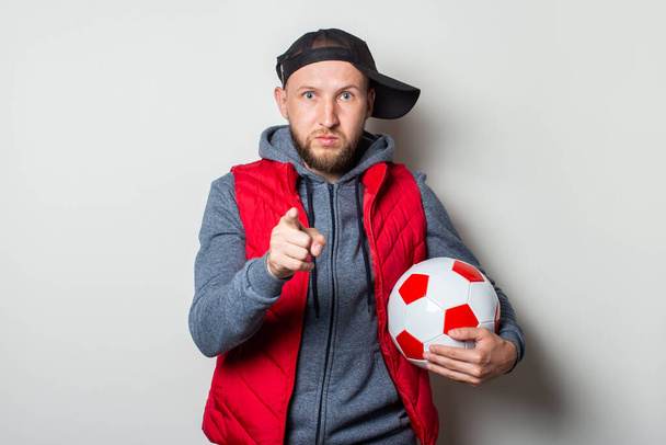 Νεαρός άνδρας με καπέλο, κουκούλα και γιλέκο κρατά μια μπάλα ποδοσφαίρου και δείχνει το δάχτυλό του στο θεατή σε ένα ελαφρύ φόντο. Είσαι η επόμενη χειρονομία.. - Φωτογραφία, εικόνα