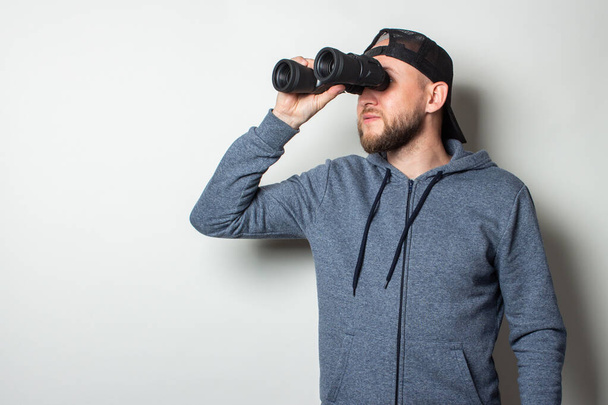 Young man in a hoodie and a cap looks through binoculars on a light background. - Foto, Imagem