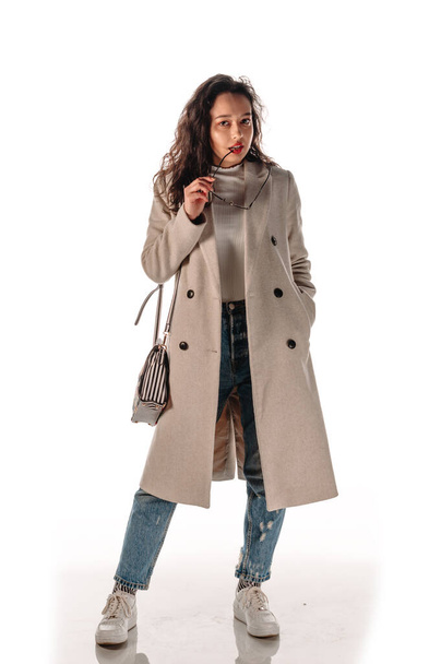 Foto de cuerpo de una chica moderna y fresca con abrigo beige y zapatillas deportivas sobre fondo blanco - Foto, imagen