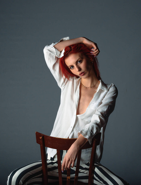 Elegante jovem mulher vestindo calças de listra e camisa branca enquanto sentado no cheir no estúdio moderno - Foto, Imagem