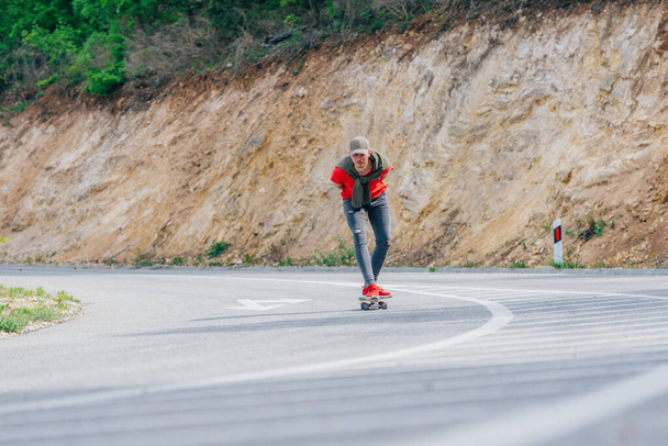 Hochgewachsener männlicher Longboarder, der an einem sonnigen Tag hoch in der Natur mit seinem Longboard bergab fährt und dabei ein rotes Hemd, eine grüne Mütze und graue Jeans trägt. - Foto, Bild