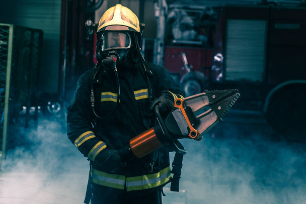 Portret van een dappere brandweerman vol vertrouwen met beschermende uitrusting, opkomst en helm. Donkere achtergrond met rook en blauw licht. - Foto, afbeelding