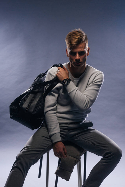 Informally ( casual ) dressed blonde model wearing a white sweater and holding a black man bag is posing on a white background while sitting on a chair. - Photo, Image