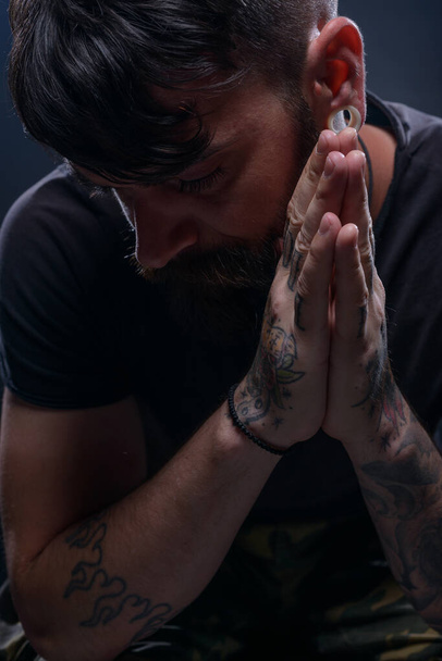 Primeros planos imagen oscura de un modelo masculino barbudo en camiseta negra sentado en una silla y rezando con sus manos tatuadas - Foto, Imagen