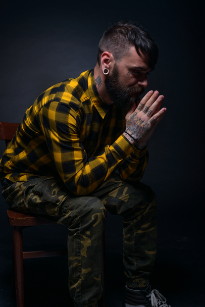 Close up portait de um modelo masculino com tatuagens e camisa de tartan sentado em uma cadeira isolada em um fundo preto - Foto, Imagem