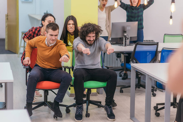 Κτίριο ομάδας και διασκέδαση γραφείου.Τέσσερις νέοι πρόσχαροι επιχειρηματίες σε smart casual φορούν διασκεδάζοντας ενώ τρέχουν σε καρέκλες γραφείου και χαμογελώντας. - Φωτογραφία, εικόνα