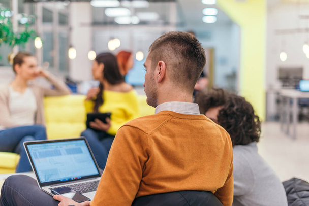 Wieloetniczni koledzy blond biały i latynoski ciemnowłosy przystojny biznesmen (pracownik) siedzi na żółtej kanapie podczas pracy na laptopie w nowoczesnej firmie startup - Zdjęcie, obraz