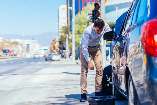 Un joven empresario cansado sudoroso se limpia el sudor de la frente mientras intenta cambiar un neumático pinchado en un hermoso día soleado. - Foto, Imagen