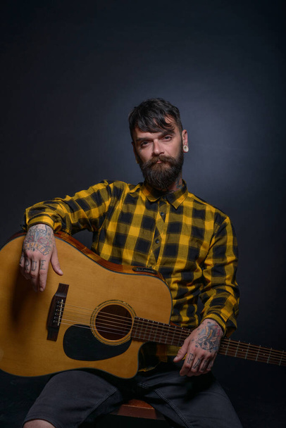 L'uomo con tatuaggi e barba sta zoccolando una chitarra e indossa una camicia nera giallo tartan - Foto, immagini
