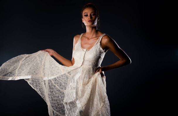 Attraktives weibliches Model in elegantem weißen Kleid posiert vor dunkler Wand auf Hintergrund - Foto, Bild