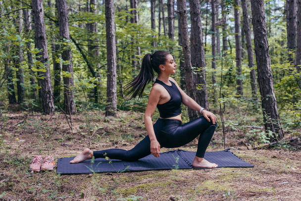 Здоровая молодая спортивная женщина делает расколы на коврике для йоги в лесу - Фото, изображение
