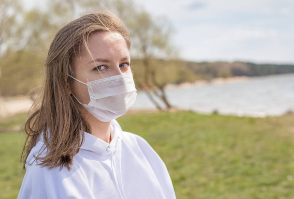 Girl, young woman in protective sterile medical mask on her face looking at camera outdoors close up. Coronavirus pandemic. Beautiful girl on the lake with medical mask. - 写真・画像