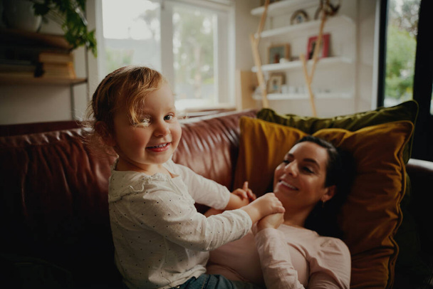 Portrait of excited little child playing with mother lying on couch at home during covid-19 lockdown - Fotografie, Obrázek