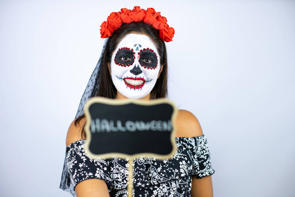 Woman wearing day of the dead costume over isolated white background smiling and holding blackboard with halloween word message - Photo, Image
