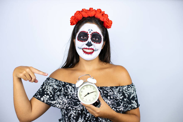 Ölü kostümü giymiş bir kadın izole edilmiş beyaz arka planda bir saat tutuyor, gülümsüyor ve onu işaret ediyor. - Fotoğraf, Görsel