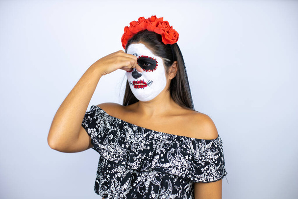 Žena v den mrtvého kostýmu na izolovaném bílém pozadí cítí něco smradlavého a odporného, nesnesitelného, zadržuje dech s prsty na nose - Fotografie, Obrázek