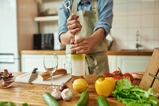 Cropped shot of young man, Italian cook opening a bottle of white wine with a corkscrew in the kitchen - Foto, imagen