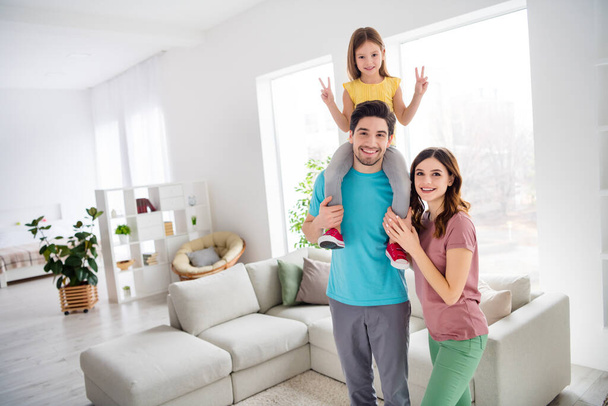 Photo of positive three people mom dad carry shoulders small girl kid make v-sign in house indoors - 写真・画像