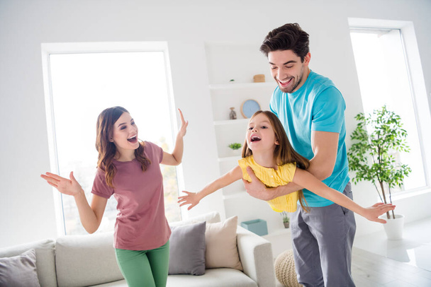 Photo of positive cheerful three people play game dad carry small kid girl she hold hands fly like bird mom show support teach how in house indoors - Fotografie, Obrázek