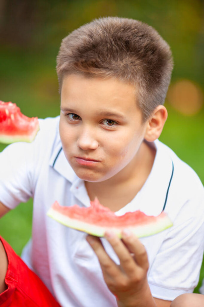 Beyaz tişörtlü çocuk parktaki çimlerde karpuz yiyor. - Fotoğraf, Görsel
