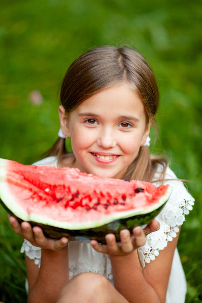 meisje in witte kleren en met twee staarten op haar hoofd eet watermeloen op het gras in het park - Foto, afbeelding