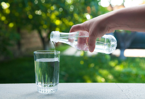 緑の自然を背景にガラスに水を注ぐハンド保持飲料水ボトル. - 写真・画像