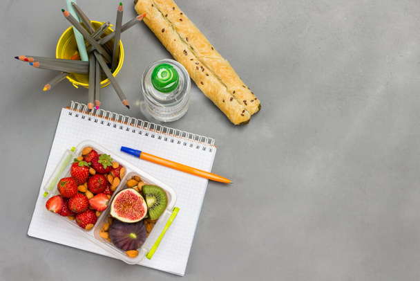 Nutritious lunch box of fruits, berries and nuts on open notebook. Bottle of water, pie and bucket of pencils. Grey background. Flat lay. Copy space - Valokuva, kuva