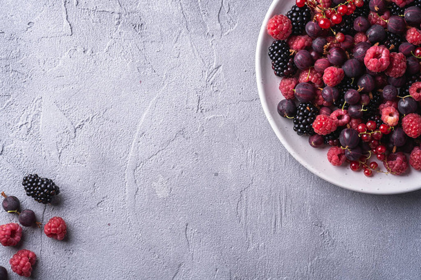 Tasty fresh ripe raspberry, blackberry, gooseberry and red currant berries in plate, healthy food fruit on stone concrete background, top view copy space - 写真・画像