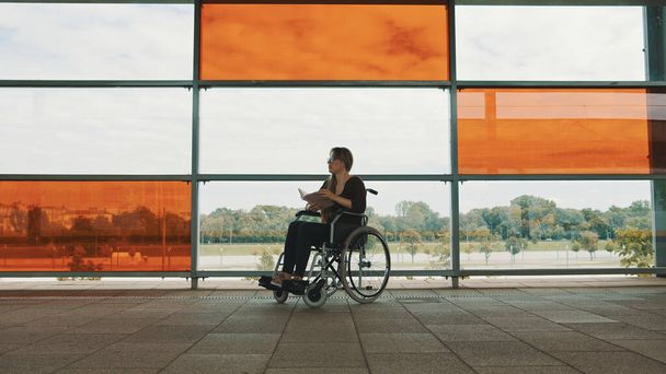 Joven mujer caucásica independiente en silla de ruedas leyendo un libro delante de grandes ventanas multicolores de cristal - Foto, imagen