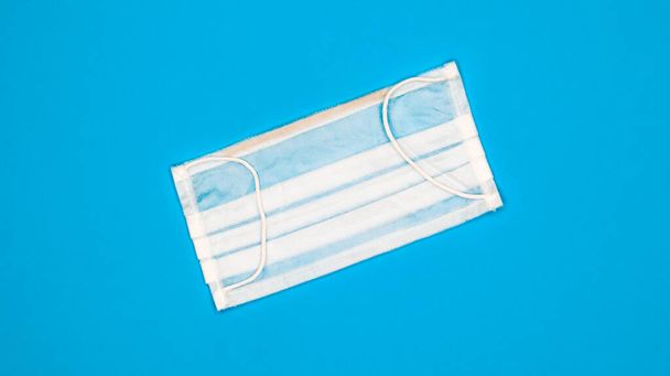 One 3-ply disposable surgical face mask with rubber grips covering mouth and nose against blue background. Protection from bacteria, healthcare and medicine concept. COVID-19. A dangerous virus - Photo, Image