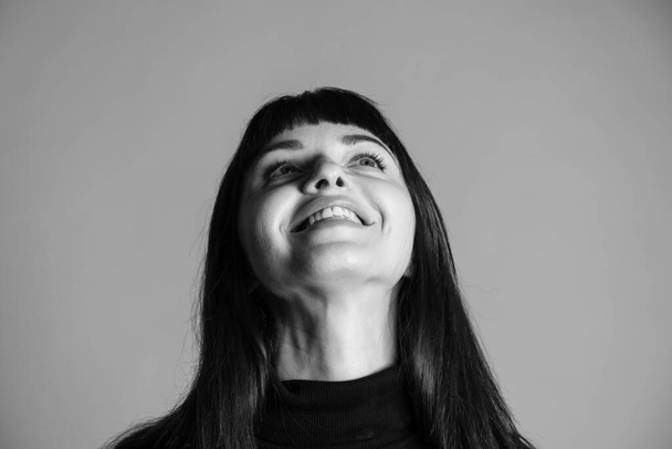 Крупный студийный портрет красивой брюнетки в сложенном черном свитере с поло-шеей, смеющейся, смотрящей в сторону, на обычном сером фоне - Фото, изображение