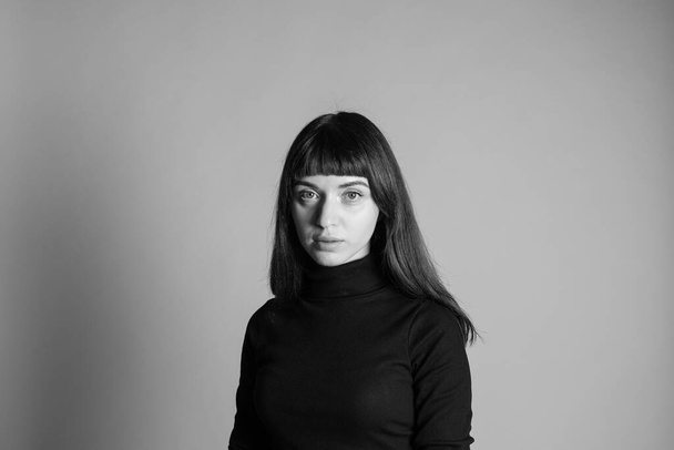 Studioporträt einer hübschen brünetten Frau, die einen gefalteten schwarzen Polohals-Pullover trägt und in die Kamera blickt, vor einem schlichten grauen Hintergrund - Foto, Bild
