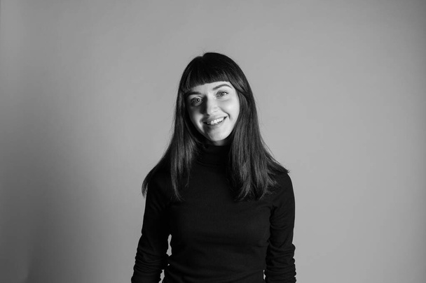 Studioporträt einer hübschen brünetten Frau, die einen gefalteten schwarzen Polohals-Pullover trägt und in die Kamera blickt, vor einem schlichten grauen Hintergrund - Foto, Bild