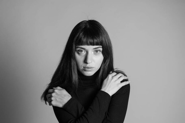 Nahaufnahme des Studioporträts einer hübschen brünetten Frau, die einen gefalteten schwarzen Polohals-Pullover trägt, die Hände auf den Schultern, in die Kamera blickt, vor einem schlichten grauen Hintergrund - Foto, Bild