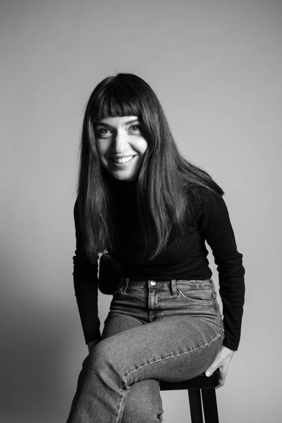 Großaufnahme des Studioporträts einer hübschen brünetten Frau, die einen gefalteten schwarzen Polohals-Pullover trägt, lacht und zur Seite schaut, vor einem schlichten grauen Hintergrund - Foto, Bild