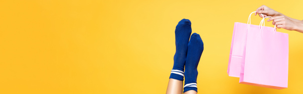Samičí nohy v modrých ponožkách v blízkosti růžových papírových sáčků izolovaných na žlutém pozadí, prapor - Fotografie, Obrázek