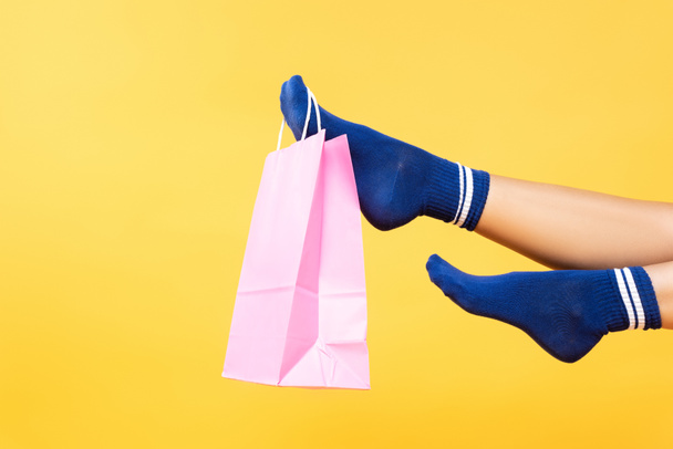 Cropped view of pink paper bag hanging on woman leg in socks isolated on yellow background - Zdjęcie, obraz