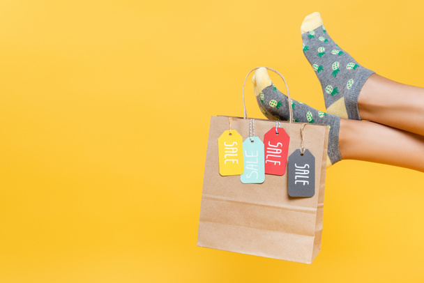 Oříznutý pohled na papírový sáček s prodejními štítky visící na ženských nohou v ponožkách izolovaných na žluté - Fotografie, Obrázek