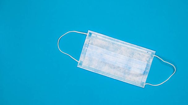 One 3-ply disposable surgical face mask with rubber grips covering mouth and nose against blue background. Protection from bacteria, healthcare and medicine concept. COVID-19. A dangerous virus - Zdjęcie, obraz