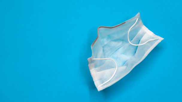 One 3-ply disposable surgical face mask with rubber pads covering the mouth and nose against a blue background. The concept of protection from bacteria, healthcare and medicine. COVID-19. copy space. - 写真・画像