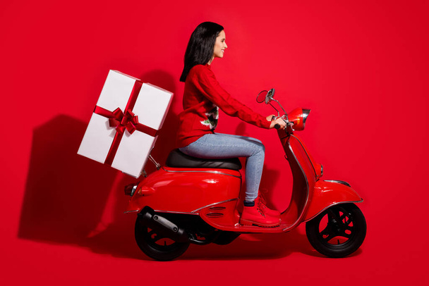 Profile side view portrait of her she nice attractive cheerful cheery girl riding bike delivering giftbox st Nicholas day fairy Santa isolated bright vivid shine vibrant red color background - Zdjęcie, obraz