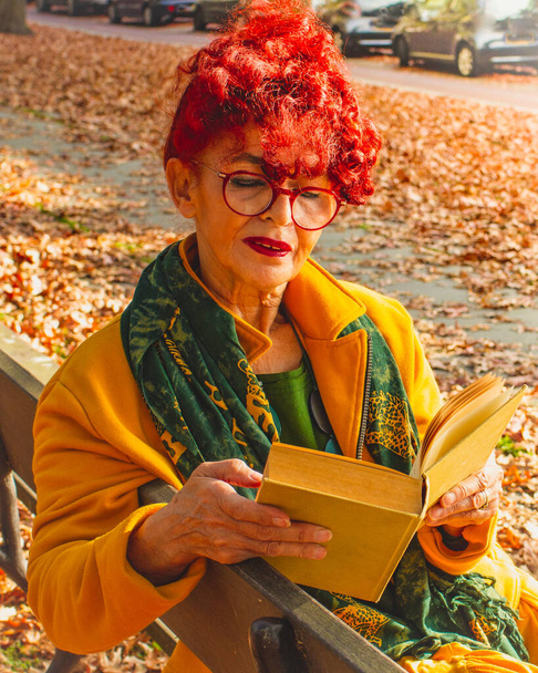 Ανώτερη γυναίκα με κόκκινα μαλλιά και γυαλιά κάθεται και χαλαρώνει σε ένα παγκάκι στην πόλη διαβάζοντας ένα βιβλίο. Σαιζόν ρούχα σε έντονα χρώματα, top view. - Φωτογραφία, εικόνα
