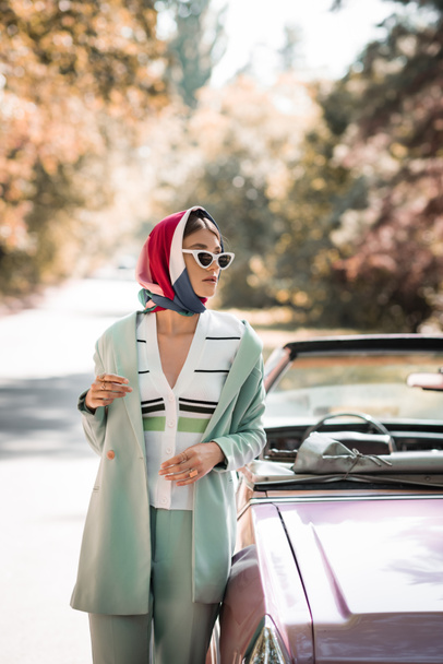 Fashionable woman in headscarf and sunglasses standing near car on road  - Zdjęcie, obraz