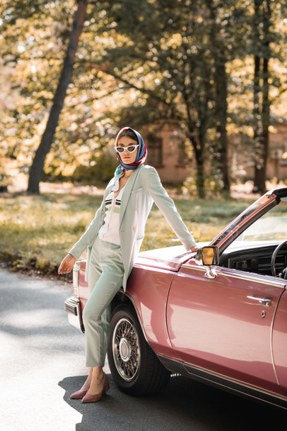 Fashionable woman in headscarf and sunglasses standing near cabriolet car on road  - Φωτογραφία, εικόνα