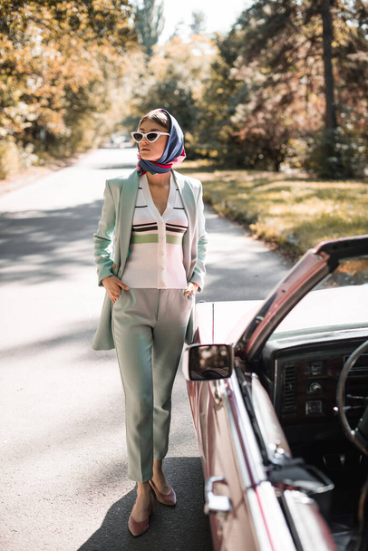 Elegant woman in sunglasses with hands in pockets looking away near car on blurred foreground on road  - Fotó, kép