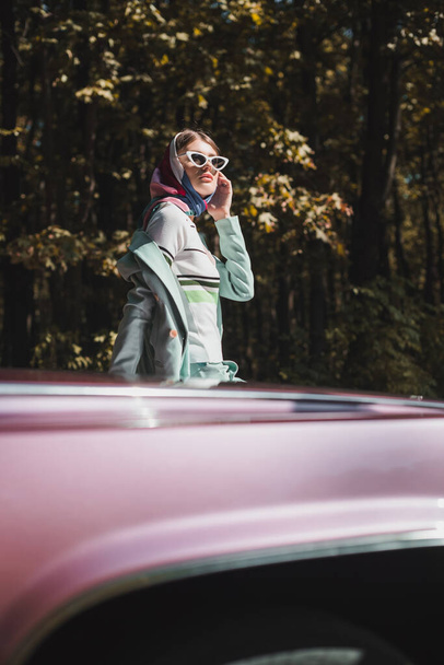 Tyylikäs nainen aurinkolaseissa katselee pois auton lähellä hämärtyneellä etualalla ulkona  - Valokuva, kuva