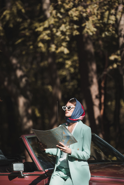 Mladá stylová žena drží mapu a dívá se pryč v blízkosti retro auto venku  - Fotografie, Obrázek