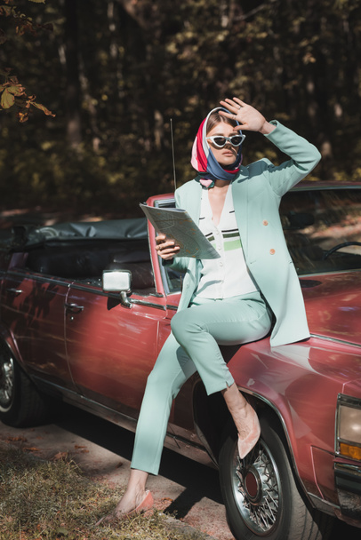 Mujer con estilo en gafas de sol sosteniendo mapa y mirando hacia otro lado cerca de coche vintage en la carretera  - Foto, imagen