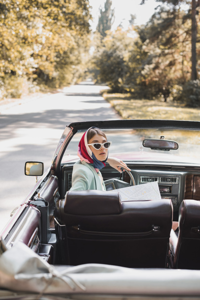 Mujer con estilo con mapa sentado en auto en primer plano borroso en la carretera durante el viaje  - Foto, Imagen